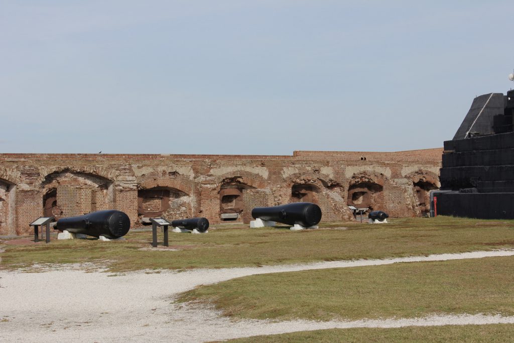 A display of the various cannon sized used in the fort with the cannon rooms in the background. The large black walls to the right are in back of a large sand bank designed to absorb a pounding from large (distant) naval guns.